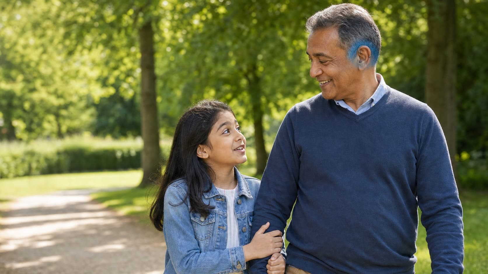 A grandfather wearing a hearing aid, smiling and walking arm-in-arm with his granddaughter in a park