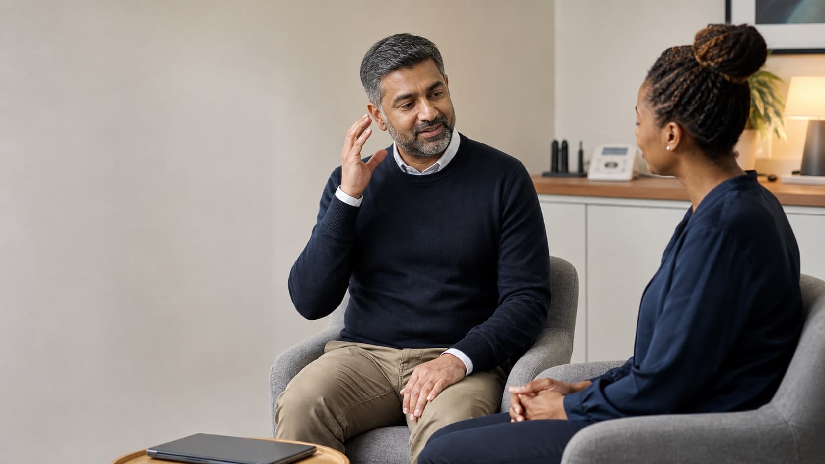 Man gesturing towards his ear while describing his symptoms to an audiologist during a consultation